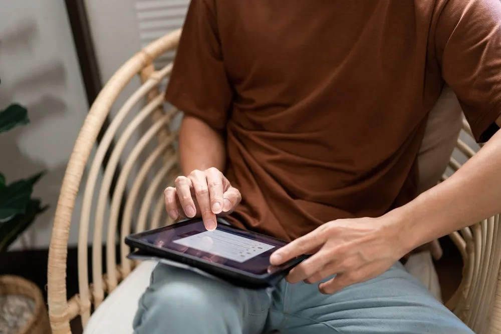 Technology Concept The male with his casual T-shirt and jeans sitting comfortably on the wooden chair and doing touchscreen for checking the web browser on the iPad.